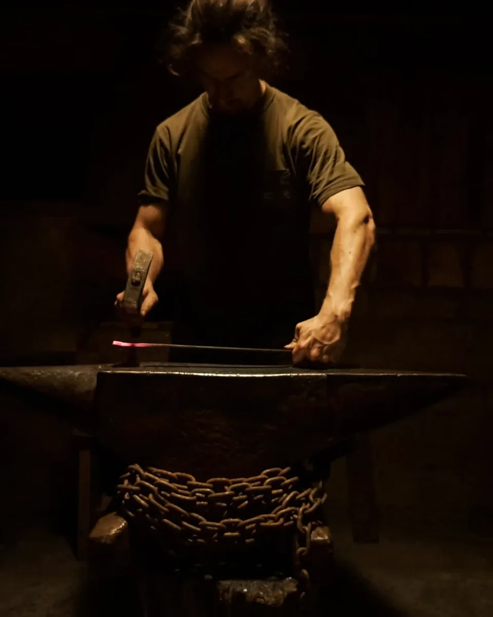 A blacksmith in a dark forge hammering a glowing steel bar on an anvil wrapped in chain